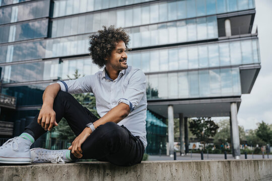 Smiling young man sitting on ledge in front of modern building