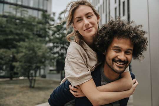 Smiling boyfriend giving piggyback ride to girlfriend in front of modern building