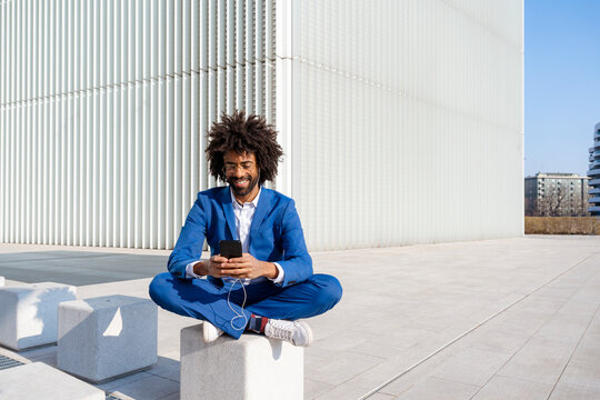 Smiling businessman listening music and using smart phone sitting on concrete block