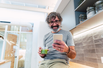 Man sitting on kitchen counter with smoothie and smart phone in kitchen at home