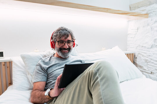 Happy senior man with headphones using tablet PC sitting on bed at home