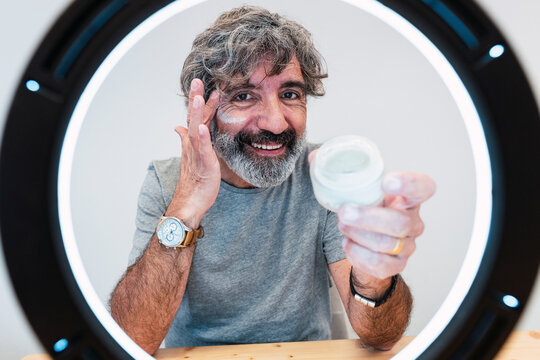 Smiling senior man applying moisturizer on face seen through ring light at home