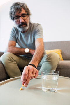 Senior Man With Glass Of Water And Picking Up Medicine Pill Sitting On Sofa At Home