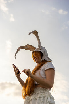 Young woman wearing rabbit costume ears using mobile phone standing under cloudy sky