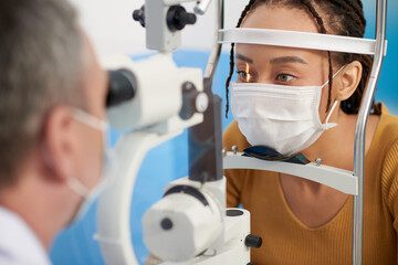 Black woman in medical mask having her eyes examined with slit lamp that allows doctor to closely...