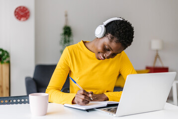 Smiling multiracial millennial female student in headphones sitting at the desk, watching webinar and making notes. Happy biracial young woman working or studying using computer at home