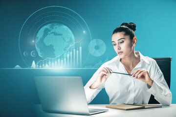 Attractive european businesswoman working at desktop with laptop and abstract glowing digital business chart and globe on blurry blue background. Trend, finance and economy concept. Double exposure.