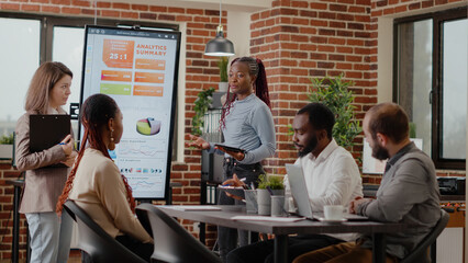 Multi ethnic colleagues presenting analysis charts on display, planning business project and strategy with people at briefing meeting. Coworkers using monitor to explain statistics.