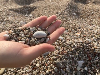 Close-up of a woman's hand. There are many multicolored stones on the palm. In the background, a beach strewn with these pebbles.