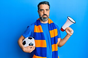 Middle age man with beard and grey hair football hooligan holding ball and using megaphone puffing cheeks with funny face. mouth inflated with air, catching air.