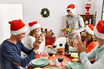 Group of people meeting clapping and sitting on the table. Man standing and holding roasted turkey celebrating Christmas at home.