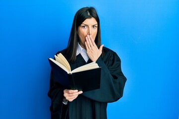 Young hispanic woman wearing judge uniform reading book covering mouth with hand, shocked and...