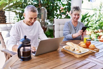 Senior caucasian couple having breakfast using laptop and smartphone at the terrace.
