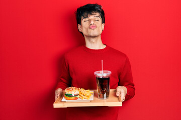 Handsome hipster young man eating a tasty classic burger with fries and soda looking at the camera...