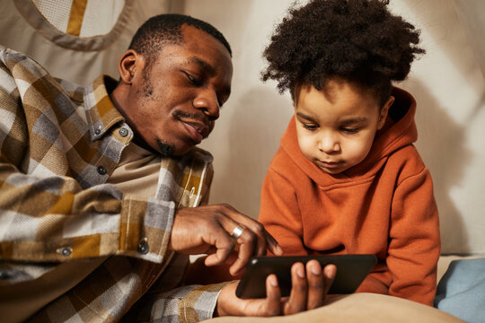 Portrait of black father with little toddler boy using smartphone together and watching cartoons in play tent