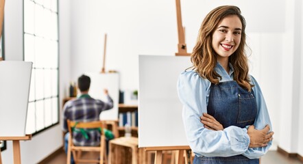 Two hispanic students painting at art school. Woman smiling happy standing with arms crossed gesture.