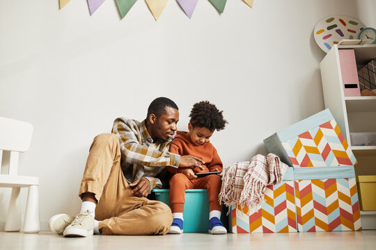 Full length portrait of African American father with little son using smartphone in cute kids room interior, copy space - Powered by Adobe