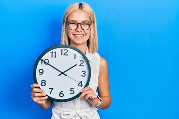 Beautiful blonde woman holding big clock looking positive and happy standing and smiling with a...