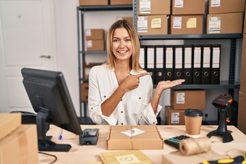 Young blonde woman working at small business ecommerce amazed and smiling to the camera while presenting with hand and pointing with finger.