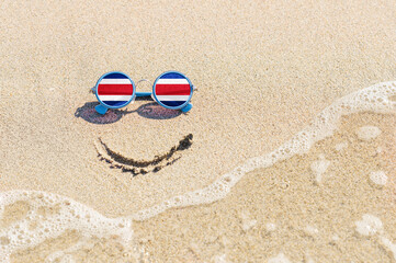 Fototapeta na wymiar Sunglasses with flag of Costa Rica on a sandy beach. Nearby is a sea lightning and a painted smile. The concept of a successful vacation in the resorts of Costa Rica.