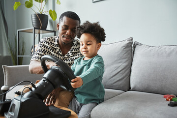 Portrait of cute black toddler playing with racing wheel with father in background, copy space