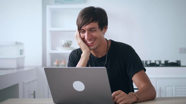 Young happy man looking at laptop monitor and laughing while reading funny news while sitting at kitchen table at home. Smiling male watches funny videos on the Internet and laughs at what he saw