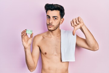 Young hispanic man shirtless wearing towel and eye bags patches with angry face, negative sign showing dislike with thumbs down, rejection concept