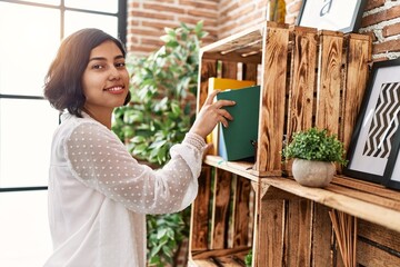 Young latin woman smiling confident holding book of shelving at home