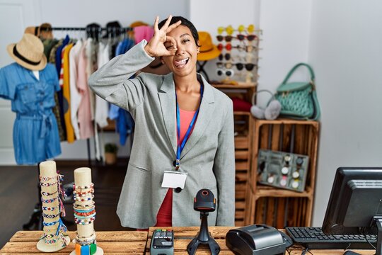 Young Hispanic Woman With Short Hair Working As Manager At Retail Boutique Doing Ok Gesture Like Binoculars Sticking Tongue Out, Eyes Looking Through Fingers. Crazy Expression.