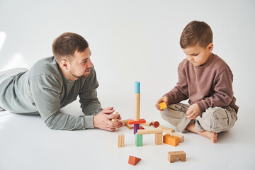 Caring dad plays with cheerful son with toy wooden cubes on white background. Fatherhood and child...