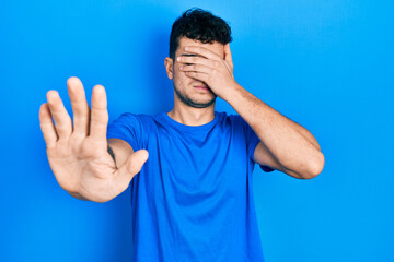 Naklejka premium Young hispanic man wearing casual blue t shirt covering eyes with hands and doing stop gesture with sad and fear expression. embarrassed and negative concept.