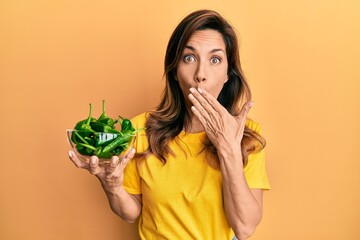 Young latin woman holding bowl with green peppers covering mouth with hand, shocked and afraid for...