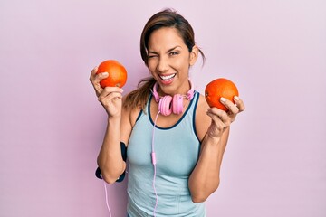 Young latin woman wearing gym clothes, using headphones and holding oranges winking looking at the camera with sexy expression, cheerful and happy face.