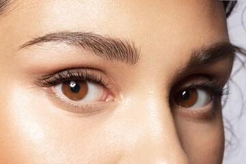 Close up of brown eyes of beautiful woman with modern makeup, shaped perfect trendy eyebrows. Female with smooth healthy skin under eye looking at camera. Make-up and skincare cosmetics advertising. - obrazy, fototapety, plakaty