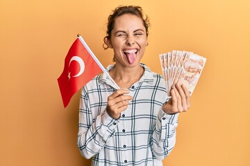 Young brunette woman holding turkey flag and liras banknotes sticking tongue out happy with funny...