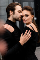 Portrait of fashion young adult couple on the photoshoot, looking directly at the camera. Handsome beard man in stylish suit hugging gorgeous woman in black dress, posing for the photo