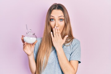 Young blonde girl holding bowl with sugar covering mouth with hand, shocked and afraid for mistake....