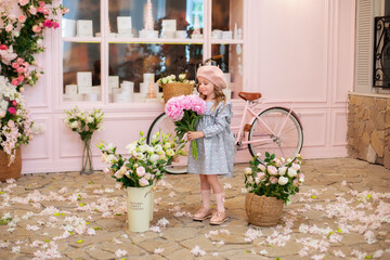 Happy little girl with curly hair in a dress and in a beret with a bouquet of pink peonies, stands...
