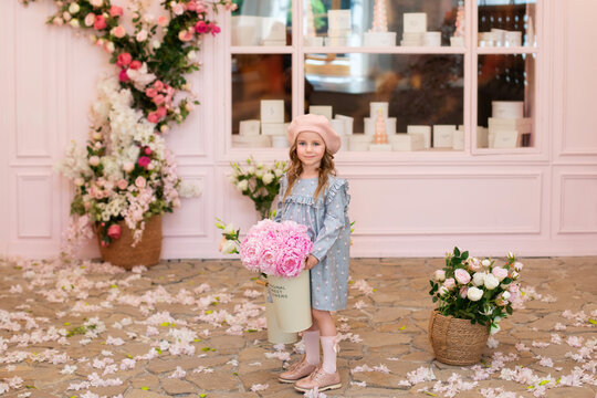 Happy little girl with curly hair in a dress and in a beret with a bouquet of pink peonies, stands near a street vintage cafe. Childhood concept. French style. Birthday. Mothers Day. Celebration.	