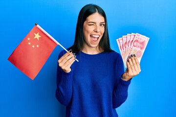 Young hispanic woman holding china flag and yuan banknotes winking looking at the camera with sexy...