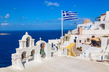 Greek orthodox church with bells and greek flag against famous white houses on Santorini island, Aegean sea, Greece.