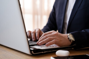 Male hands typing on laptop in office or at home on background of window in stylish modern interior.Unrecognizable businessman wearing suit and watch at work, close-up