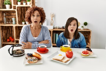 Family of mother and down syndrome daughter sitting at home eating breakfast afraid and shocked with surprise expression, fear and excited face.