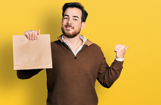Young hispanic man holding take away paper bag pointing thumb up to the side smiling happy with open mouth