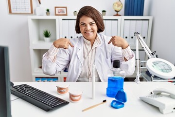 Middle age hispanic dentist woman working at medical clinic looking confident with smile on face,...
