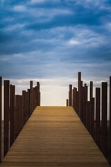 A boardwalk with vertical wood piles on both sides leading to the unknown and a dramatic sky on top. Path to light, hope or future concept. Vertical composition. Quiaios Beach, Portugal