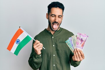 Young hispanic man holding india flag and rupee banknotes sticking tongue out happy with funny...
