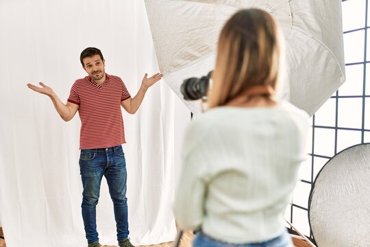 Woman photographer talking pictures of man posing as model at photography studio clueless and confused expression with arms and hands raised. doubt concept.