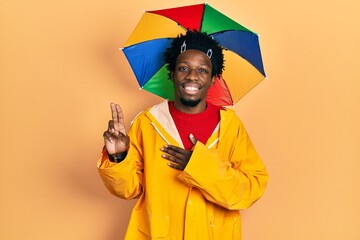 Young african american man wearing yellow raincoat smiling swearing with hand on chest and fingers...