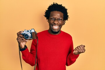 Young african american man holding vintage camera screaming proud, celebrating victory and success very excited with raised arm
